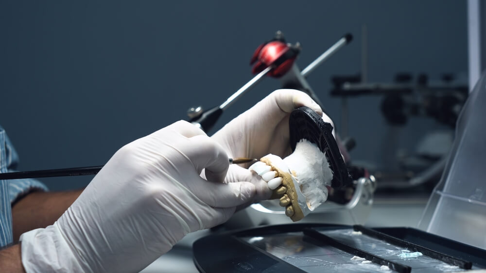 A person wearing white gloves crafts a dental model using a brush in a lab setting.