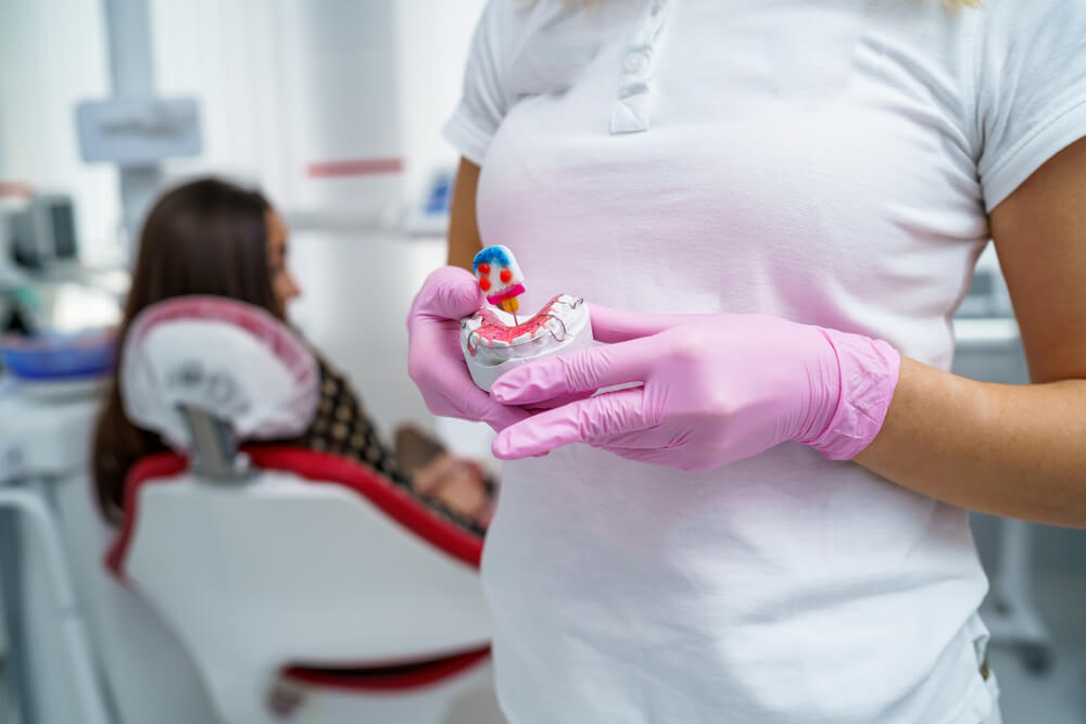 Dentist in pink gloves holds a dental mold with a small model tooth. A patient sits in a dental chair in the background.