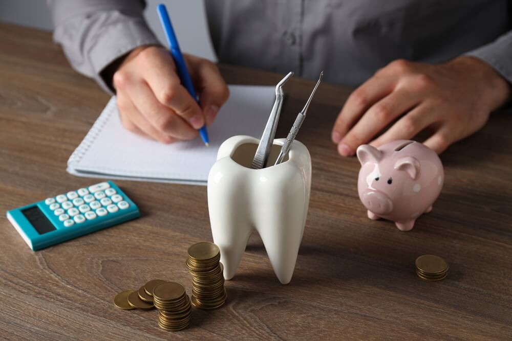 A hand writing notes next to a tooth-shaped utensil holder with dental tools, a calculator, coins, and a piggy bank on a wooden table.