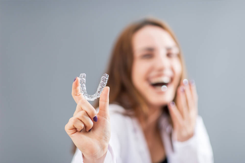 A woman smiling while holding a clear aligner, showcasing its use as a mouth guard for teeth alignment.